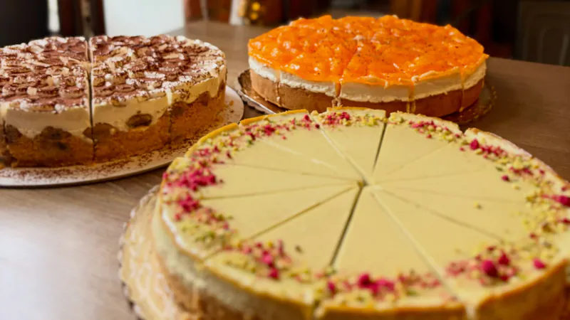 Tri torty na stole pri okne kaviarne: tiramisu cheesecake, mandarínková torta a pistáciový cheesecake s drvenými pistáciami a ružovou posýpkou